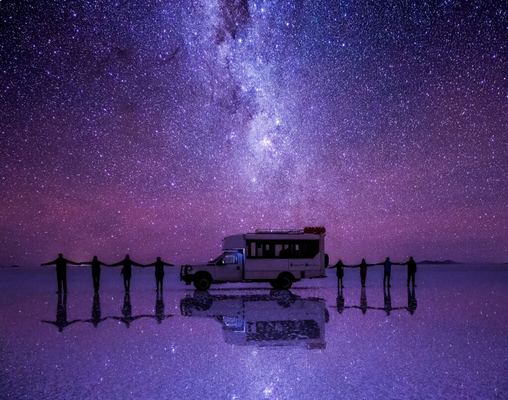 A group of guests stand arms out around a van on the reflective salt flats with the milky way trailing overhead.